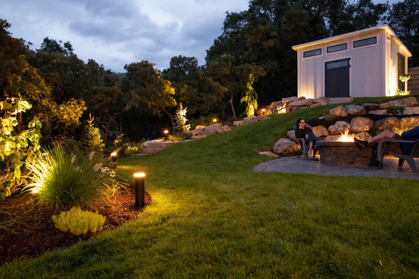 A sloped garden lit at dusk with path lights and a firepit gathering