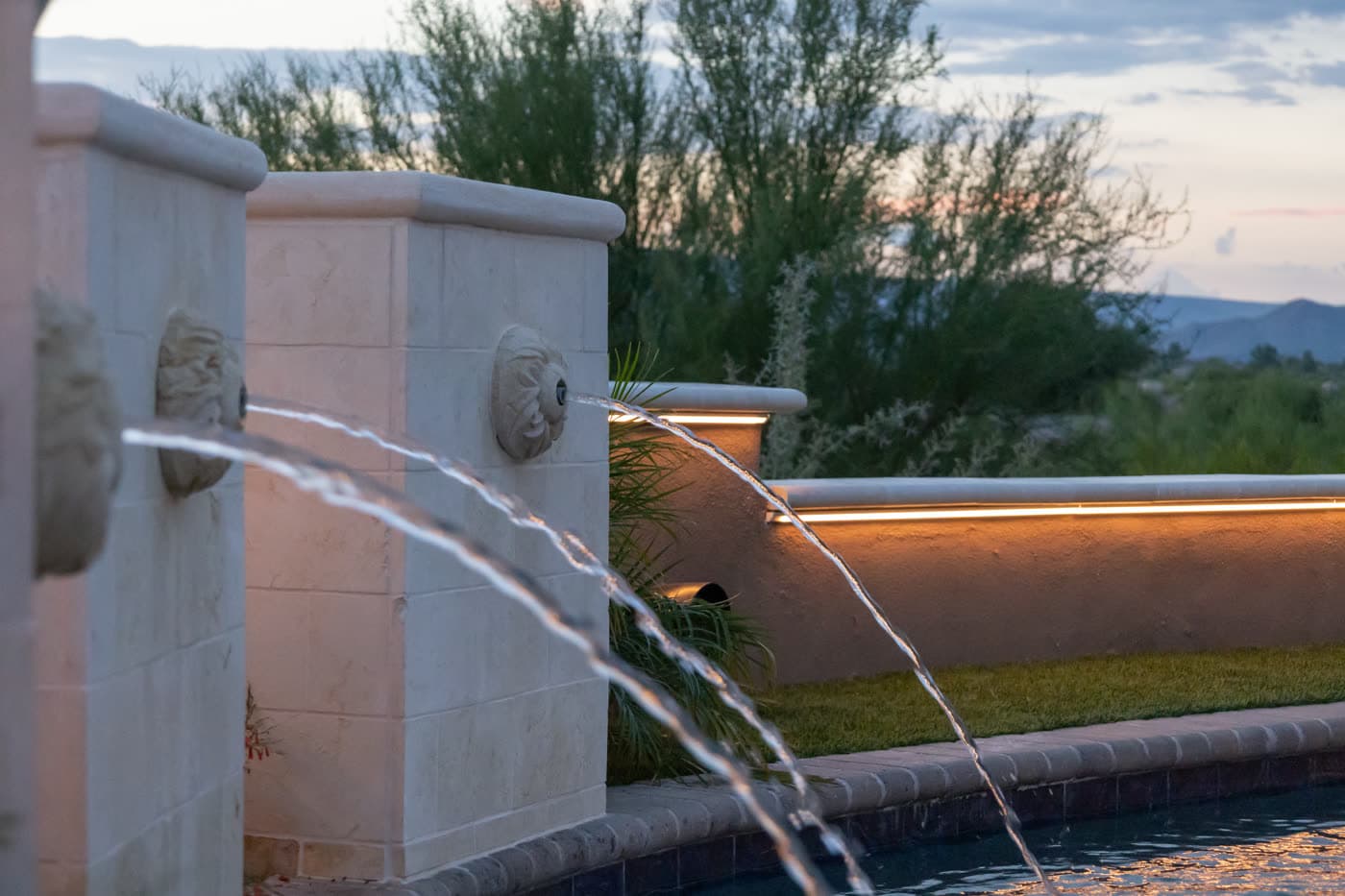 A poolside water feature lit warmly at dusk with a distant mountain view